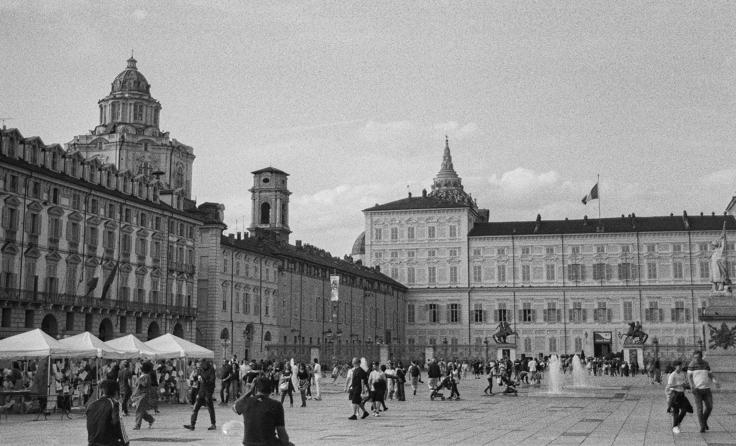 Vista della piazza con numerosi passanti e bancarelle, circondata da eleganti edifici storici.